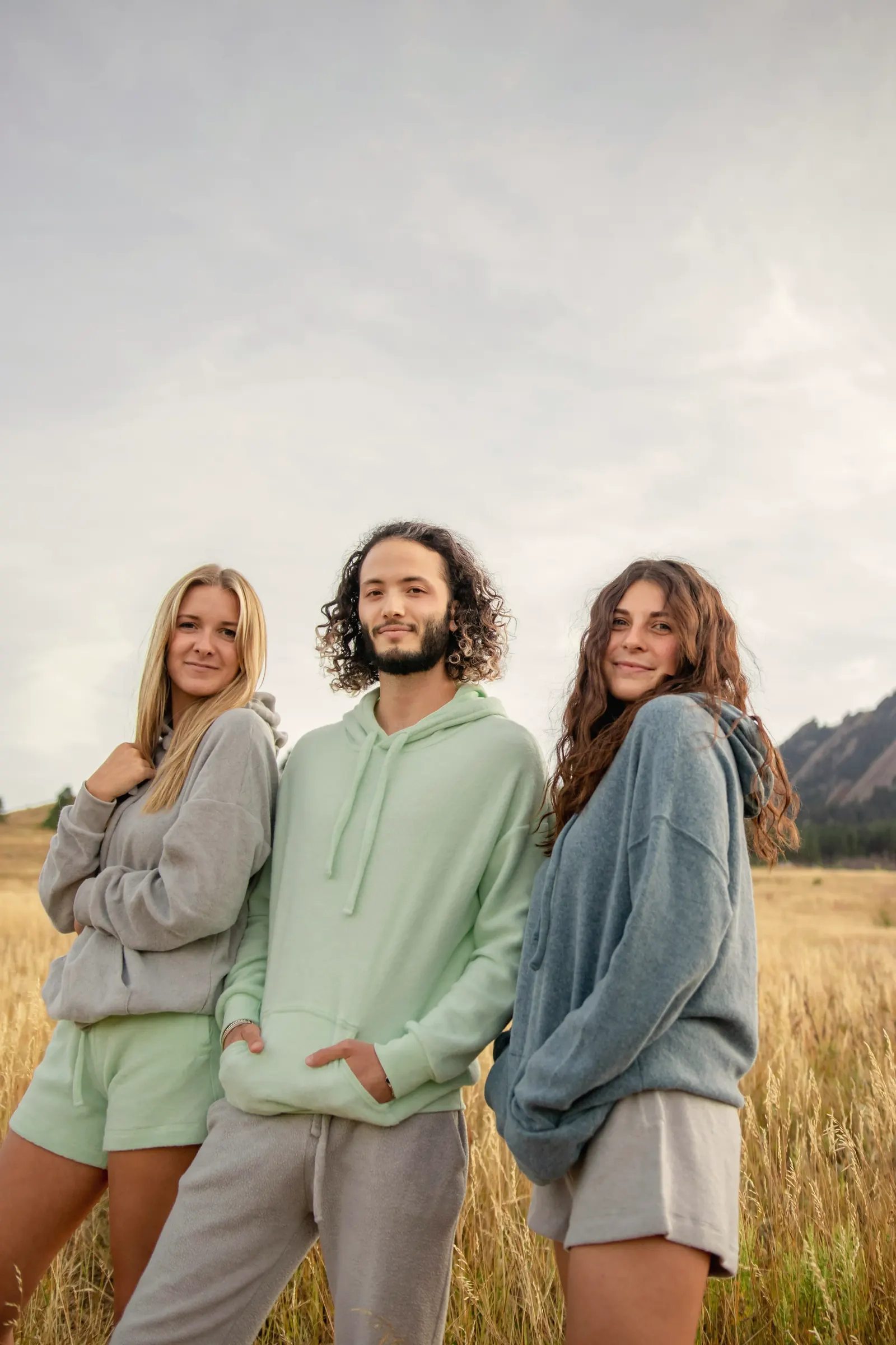 Three friends leaning into each other in a dry grass field, wearing relaxed grey, mint and slate fleece hoodies and sweats under an overcast sky